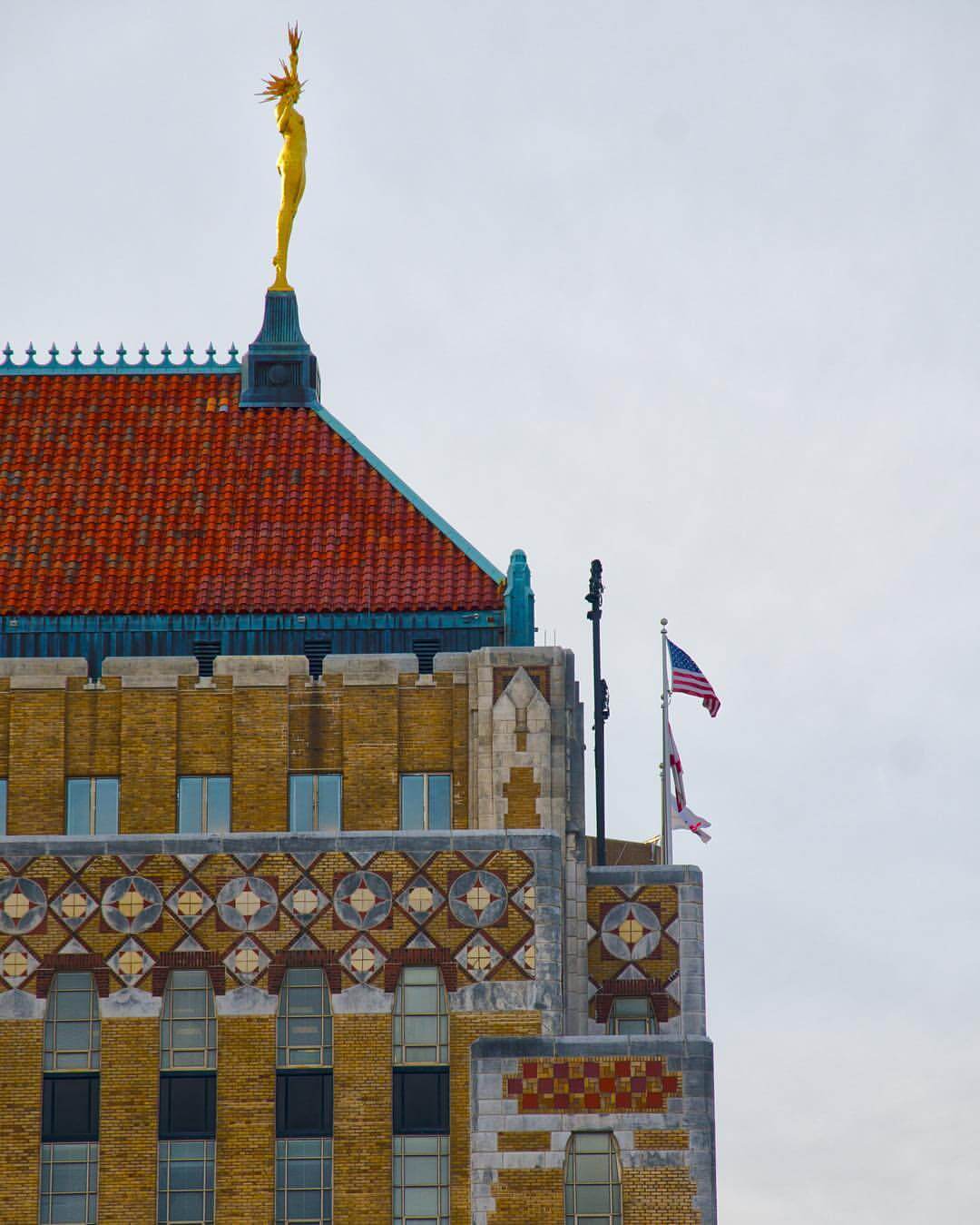 An Art Deco building graced by Miss Electra on the top Its red tiled roof complements intricate geometric patterns, while flags flutter spiritedly against a cloudy sky.