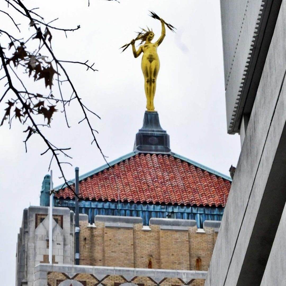 A golden statue of Birmingham's Golden Goddess, known as Miss Electra, holds lightning bolts atop a building with a tiled roof, set against a cloudy sky. Bare tree branches are visible on the left.