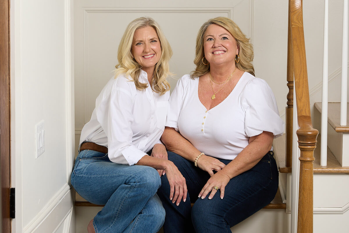 Two women, exuding clarity and confidence, are seated on stairs indoors. Both wear white shirts and jeans, beaming at the camera with the vibrant energy characteristic of ATL moms.