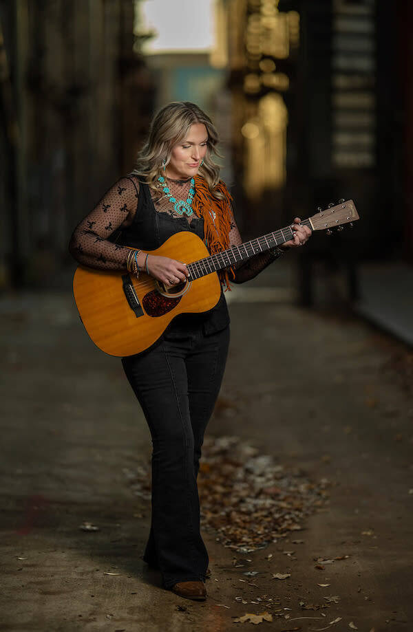 Americana artist Rebekah Gilbert, with blonde hair, plays an acoustic guitar while walking down an alleyway covered with scattered leaves.