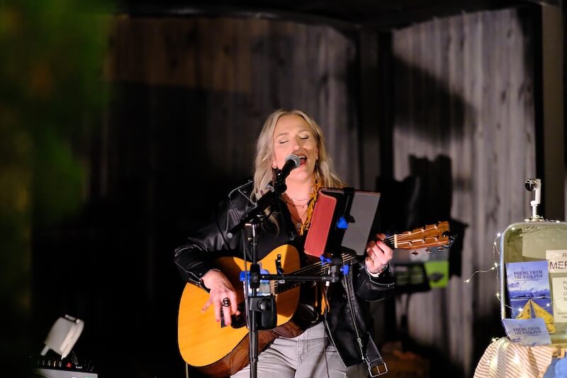 Americana artist Rebekah Gilbert, with long blonde hair, plays an acoustic guitar and sings into a microphone during a BHAM performance. A music stand and various equipment are visible in the foreground.