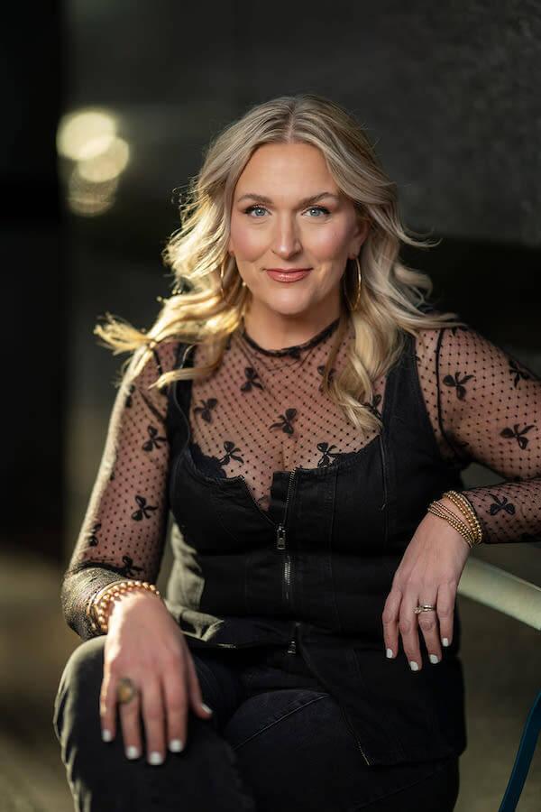 A woman with long blonde hair sits on a bench, wearing a black mesh top with butterfly designs and hoop earrings, looking directly at the camera.