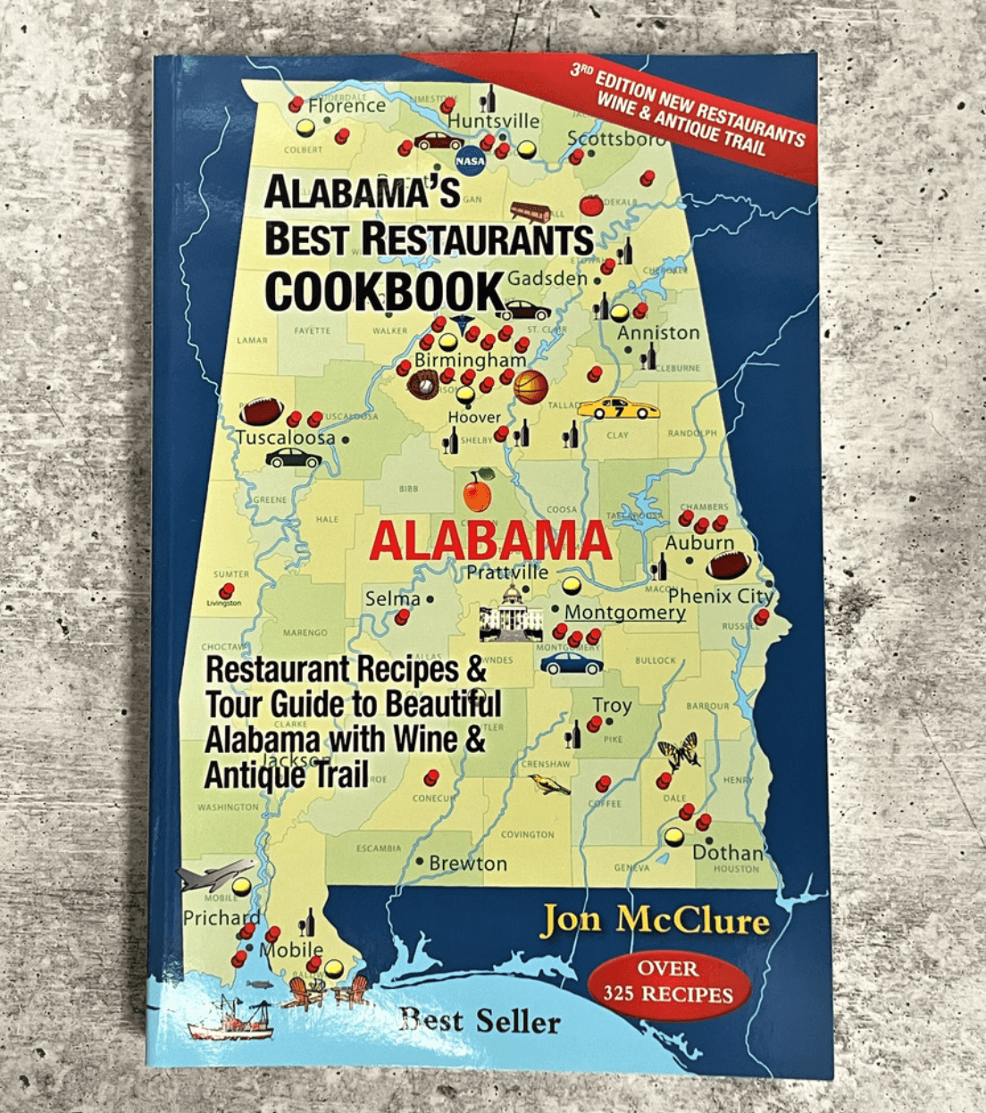 A cookbook titled "Alabama's Best Restaurants Cookbook" by Jon McClure, featuring a map with Birmingham and other cities marked, lies on a textured gray surface—perfect for discovering new May FINDS across Alabama.