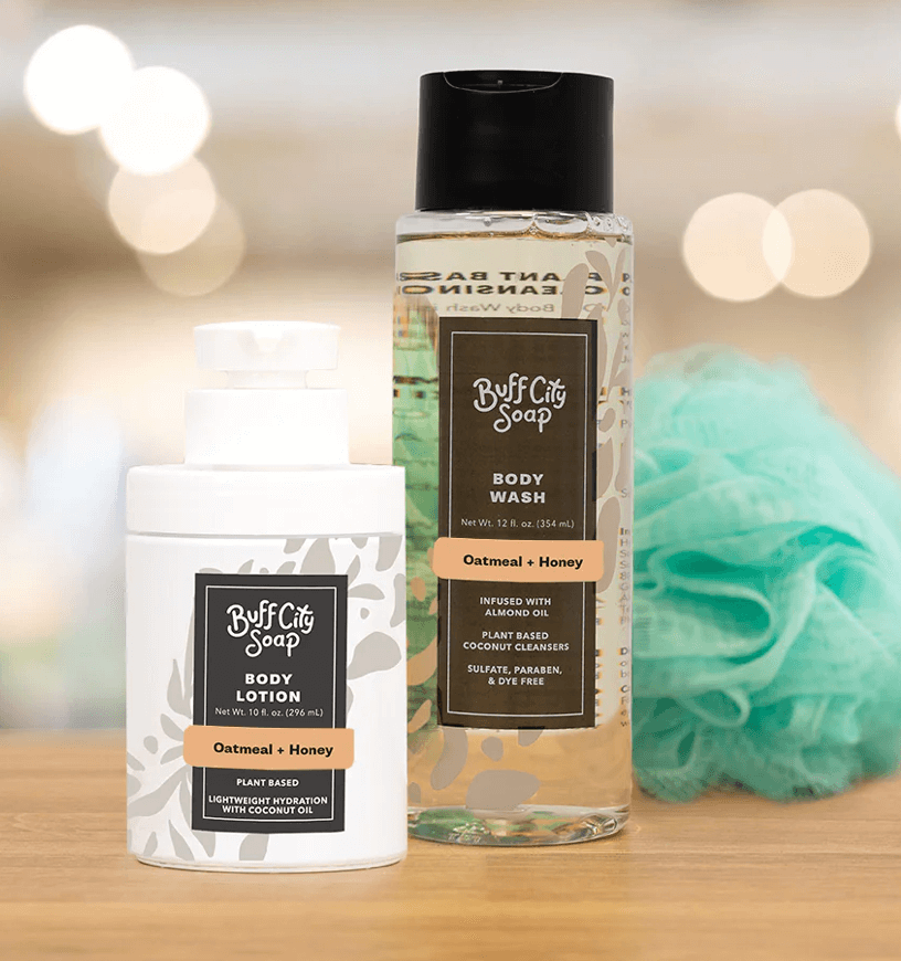 Two Buff City Soap products labeled "Oatmeal + Honey"—a body lotion and a body wash—stand next to a green bath pouf on a light surface, with a blurred background. 