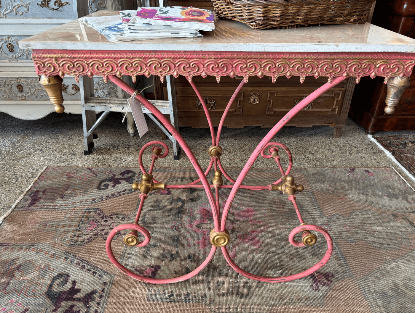 A pink wrought iron table with gold accents and a marble top, styled on a patterned rug. Magazines and a wicker basket sit atop this charmin find.