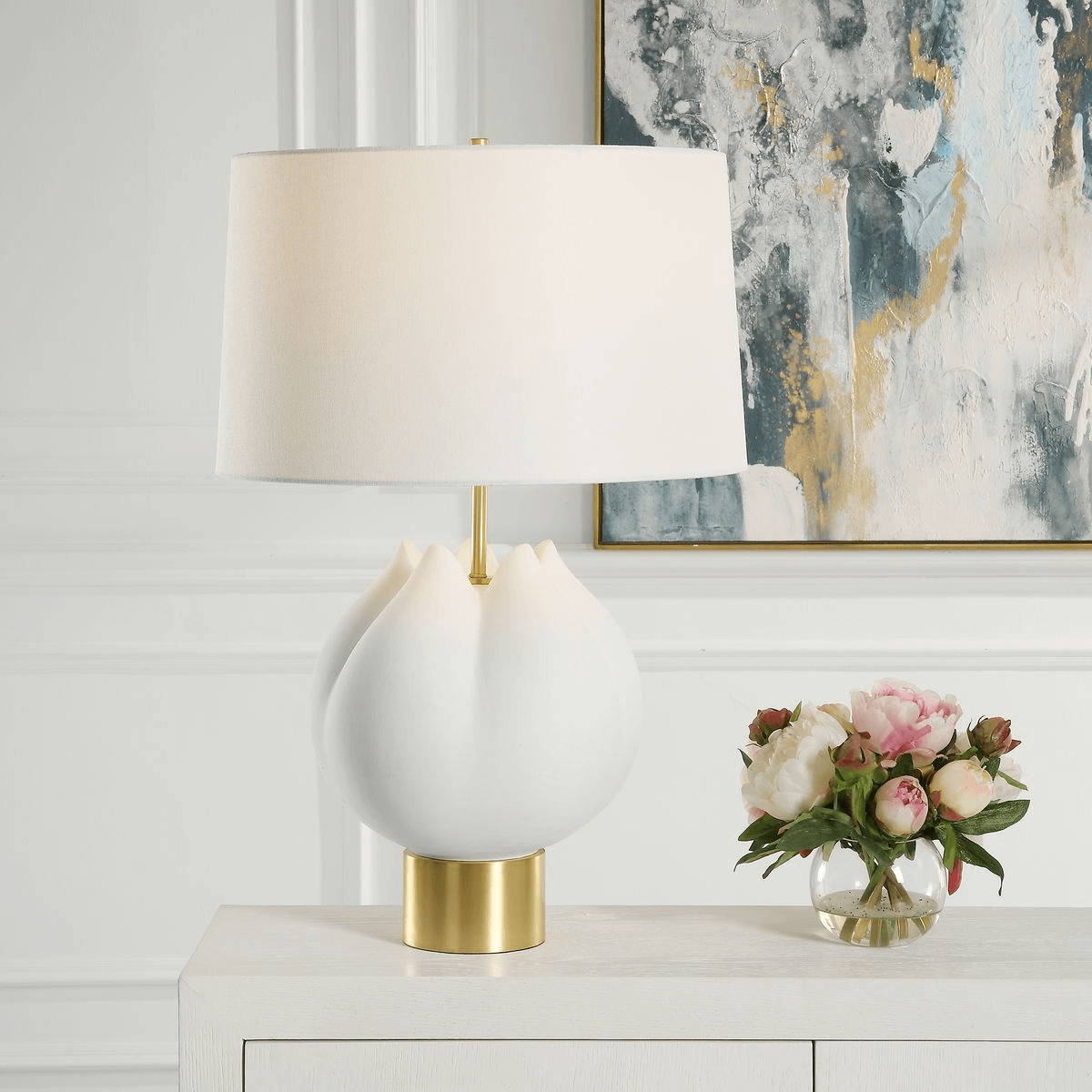 White table lamp with a sculpted base and gold accents sits on a white sideboard, paired with pink and white flowers