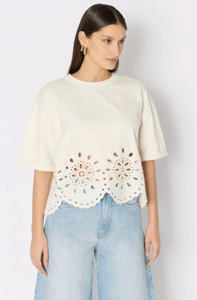A woman wearing a loose white top with floral cut-out designs along the hem, paired with light blue jeans, stands against a plain background.