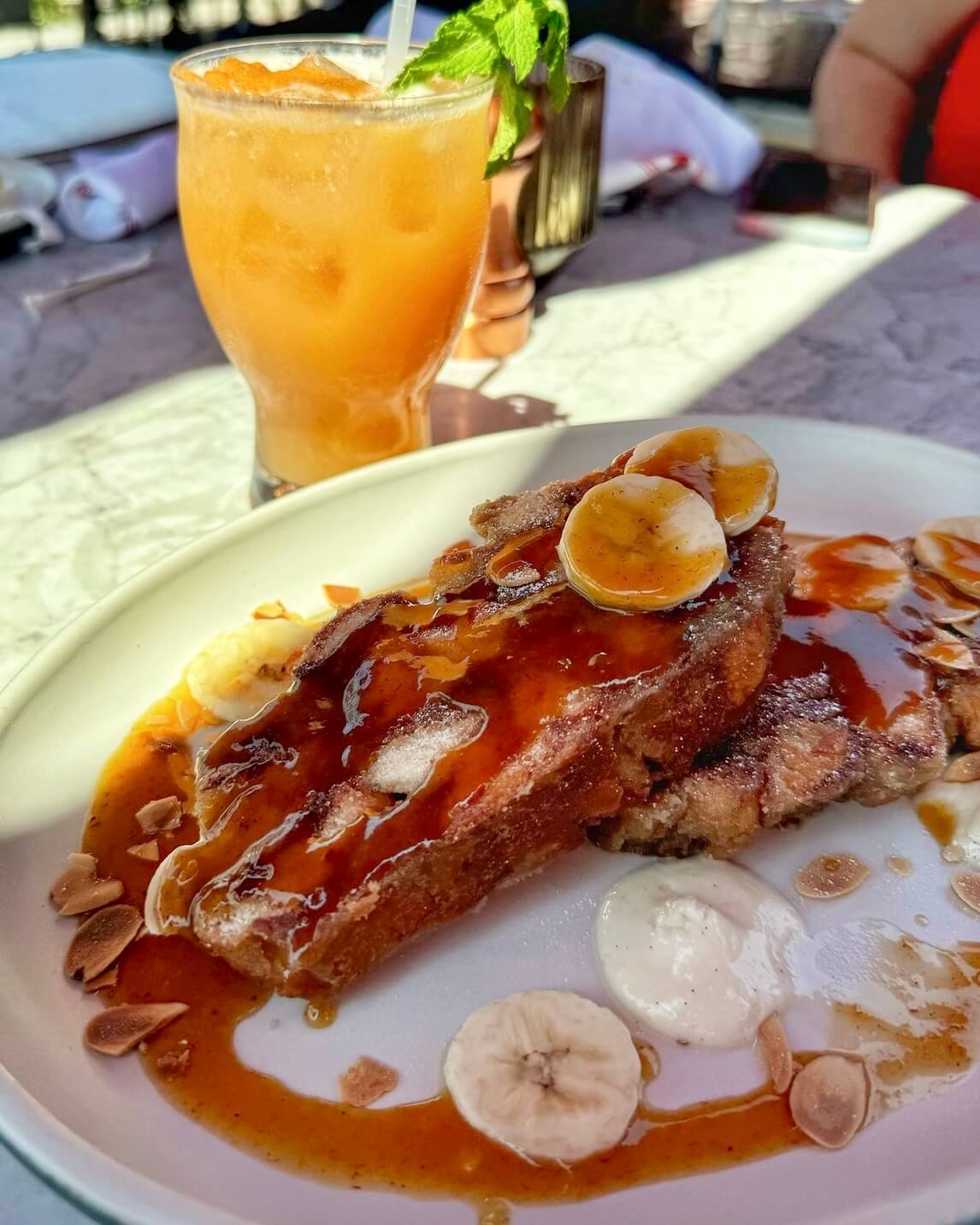 A plate of French toast topped with banana slices and syrup, garnished with nuts beside a glass of orange-colored drink—perfect for a Mother's Day brunch in Birmingham, AL.