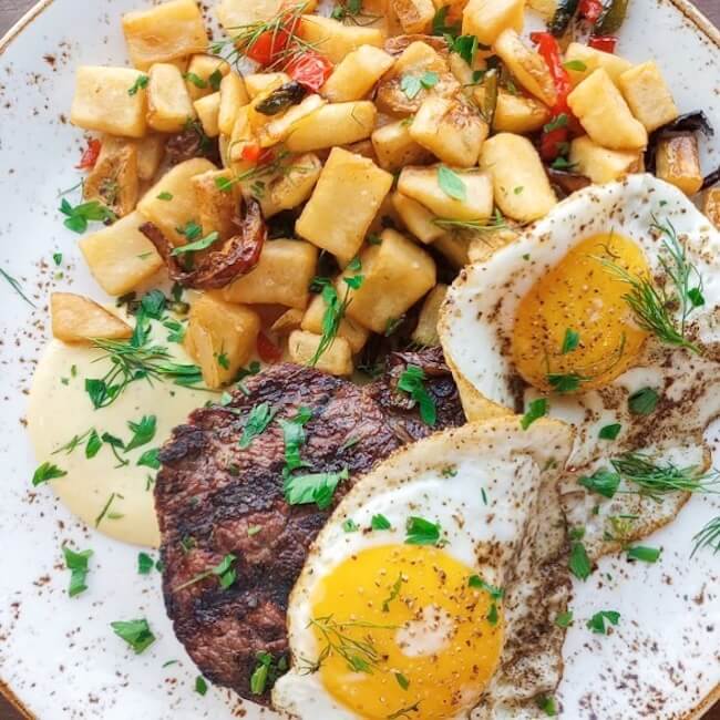 A plate with two sunny-side-up eggs, a sausage patty, seasoned potato cubes, garnished with fresh herbs and a dollop of creamy sauce—perfect for a Mother’s Day brunch in Birmingham, AL.