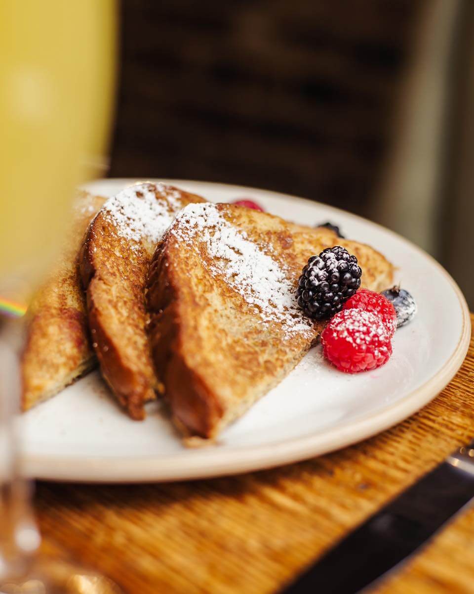 Three slices of French toast dusted with powdered sugar are served on a white plate with raspberries and a blackberry—perfect for a Mother's Day brunch in Birmingham, AL.