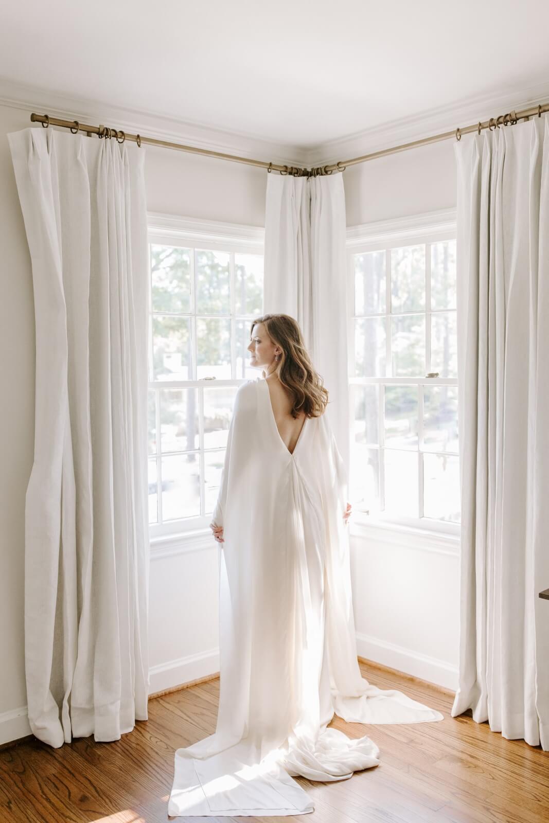 A woman in a long white wedding gown stands by two large windows in a bright room with white curtains and wooden floors.