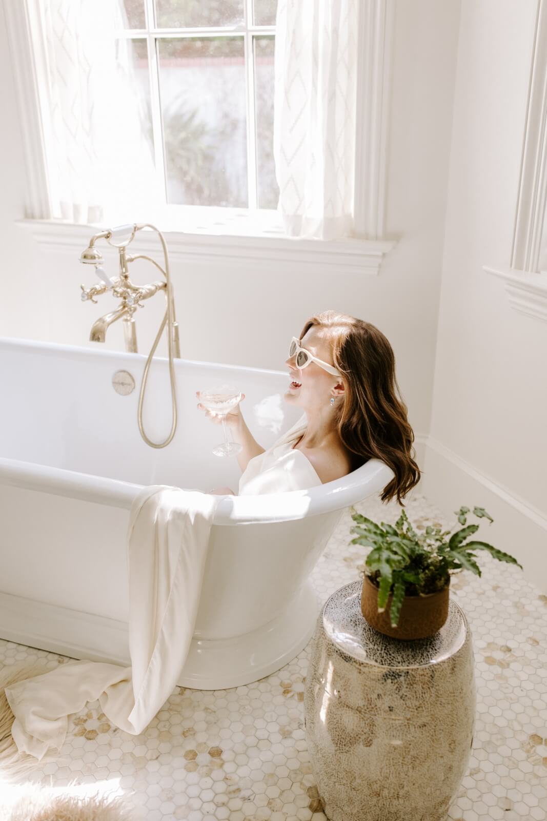 A woman wearing sunglasses relaxes in a white bathtub, holding a drink, in a bright bathroom with a window and a small plant on a stool.