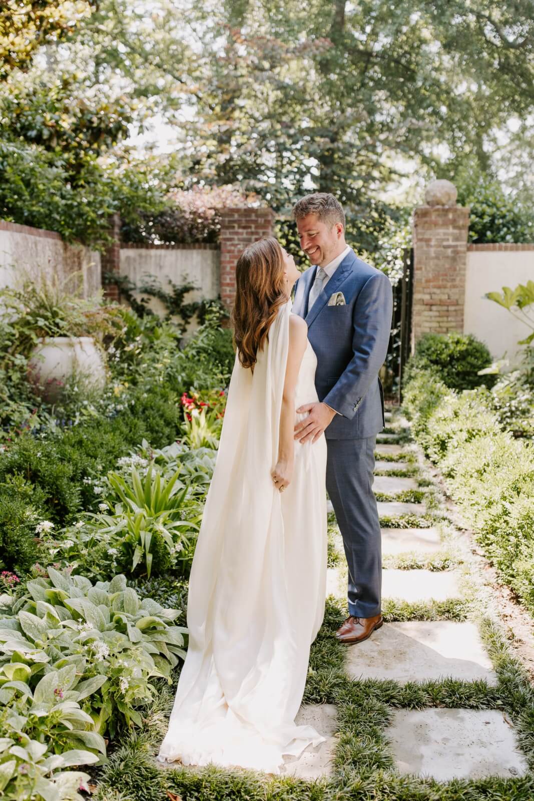 A bride in a white dress and groom in a blue suit stand smiling at each other in a lush garden, surrounded by greenery and stone pathways.