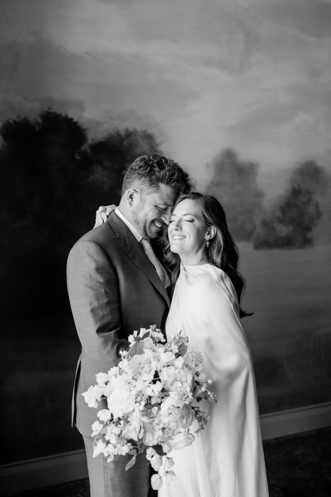 A couple in wedding attire stands closely together, smiling and embracing, with the bride holding a bouquet of flowers in front of a painted landscape backdrop at their elegant Homewood brunch wedding.