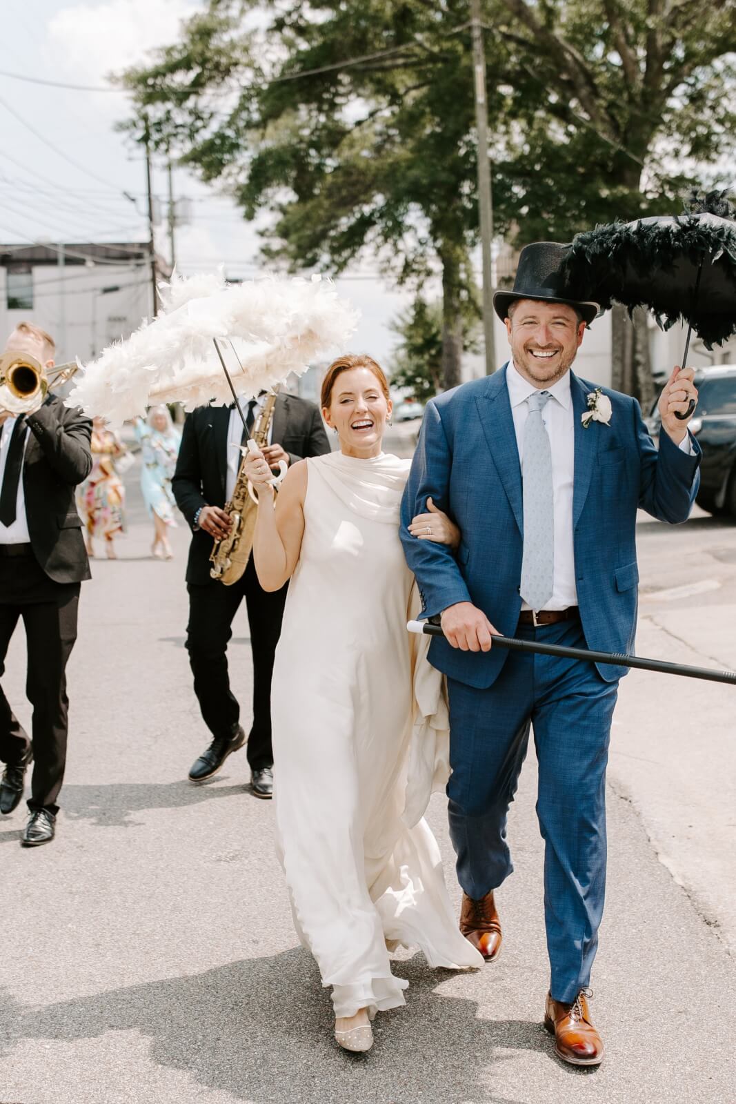 A bride and groom walk arm-in-arm down a street, smiling, holding feathered parasols in a joyful second line sendoff, followed by a band with brass instruments.