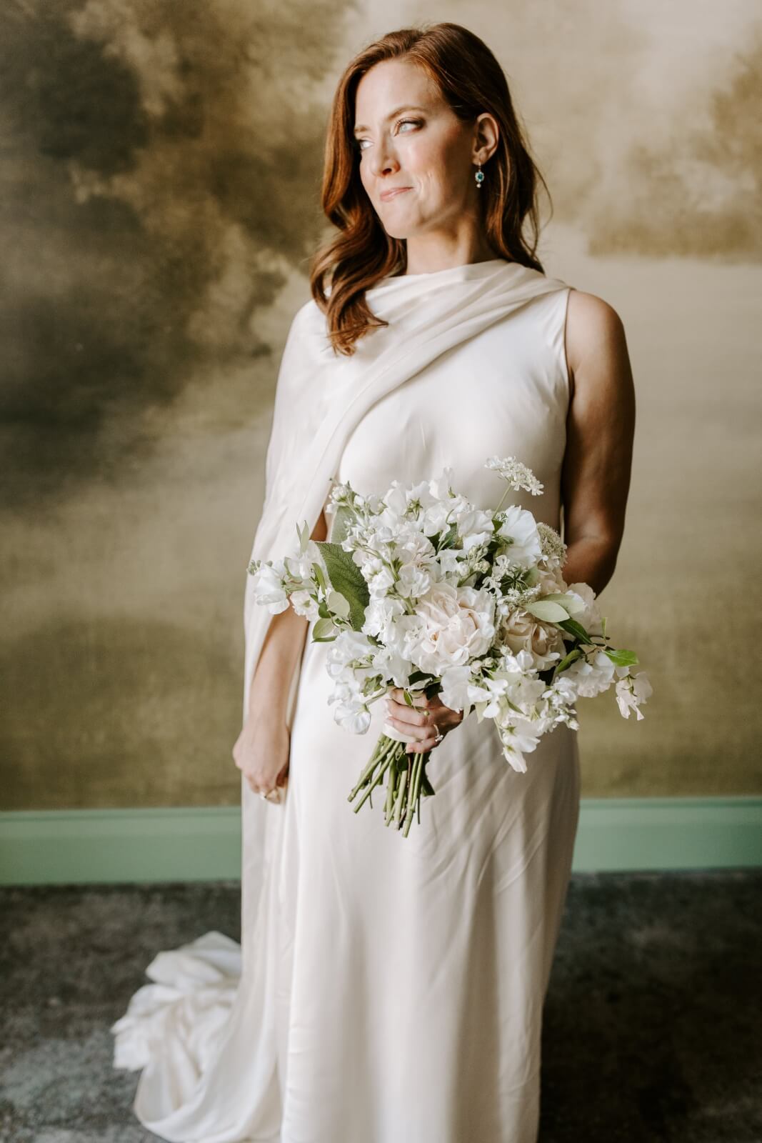 A woman in a white wedding gown holds a bouquet of white flowers, standing in front of a muraled background at her elegant Homewood brunch wedding.