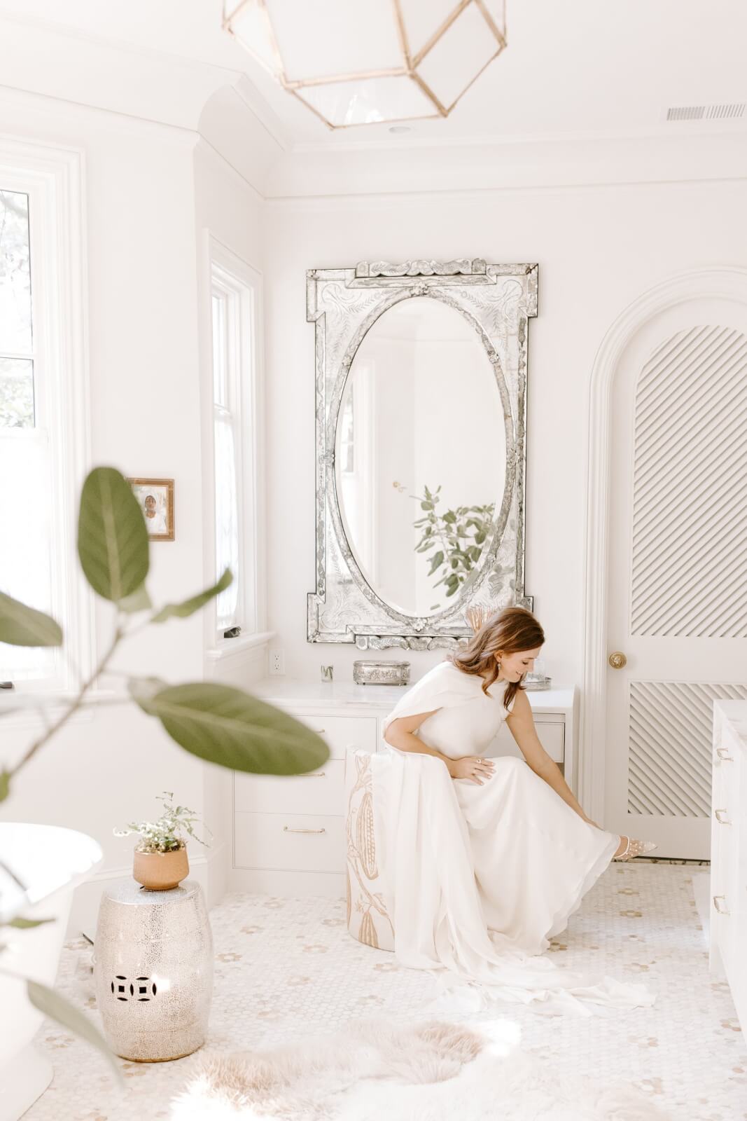 A woman in a white dress sits on a bathroom vanity adjusting her shoe. The bright space features white walls, a large ornate mirror, and potted plants.