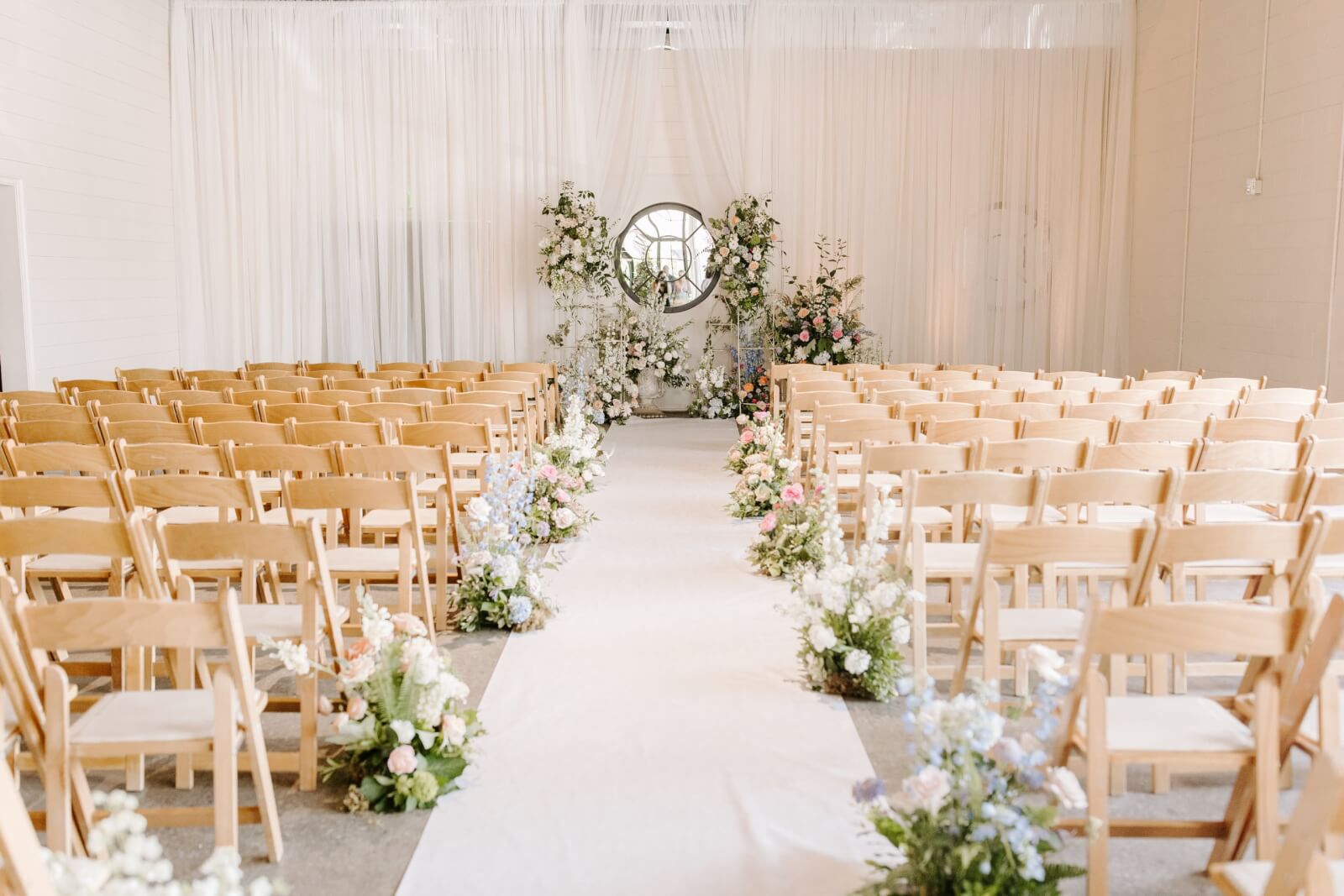 Rows of wooden chairs face a floral-decorated altar with a round mirror at the front of a white-draped indoor Homewood wedding ceremony space. The aisle is lined with flower arrangements, creating an elegant setting for a memorable celebration.