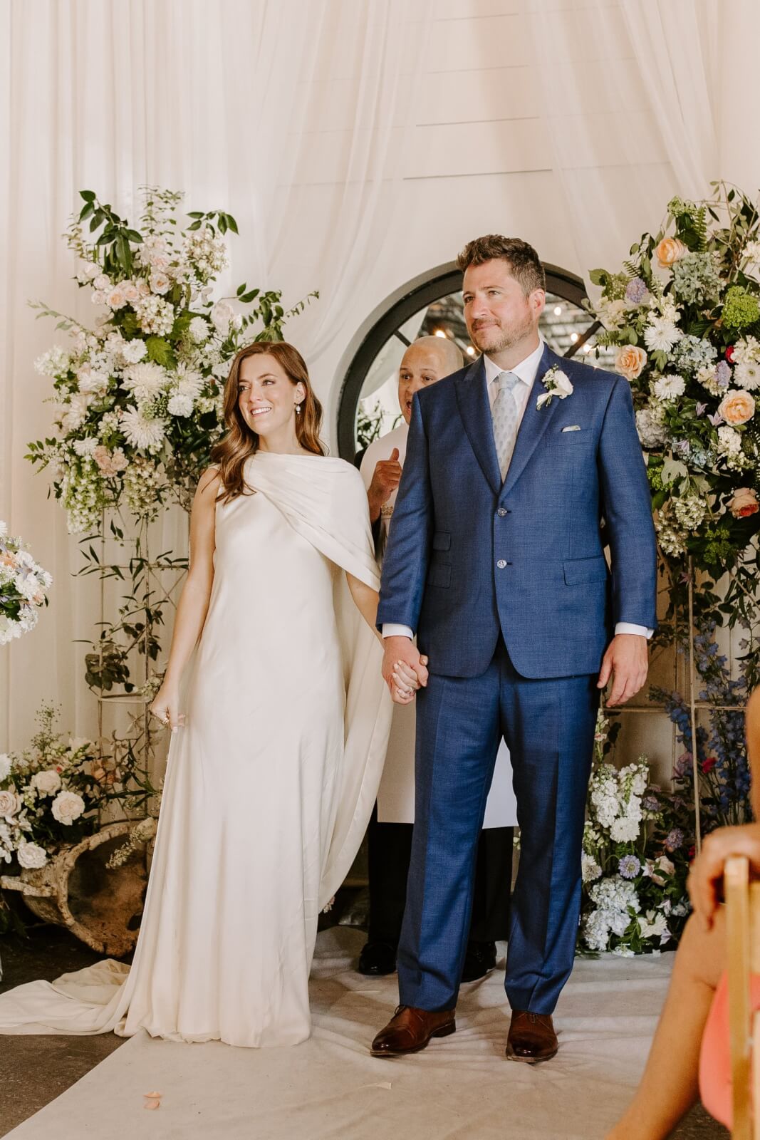 A bride in an ivory dress and a groom in a blue suit stand holding hands amid floral arrangements at the alter looking out to their gathered loved ones.