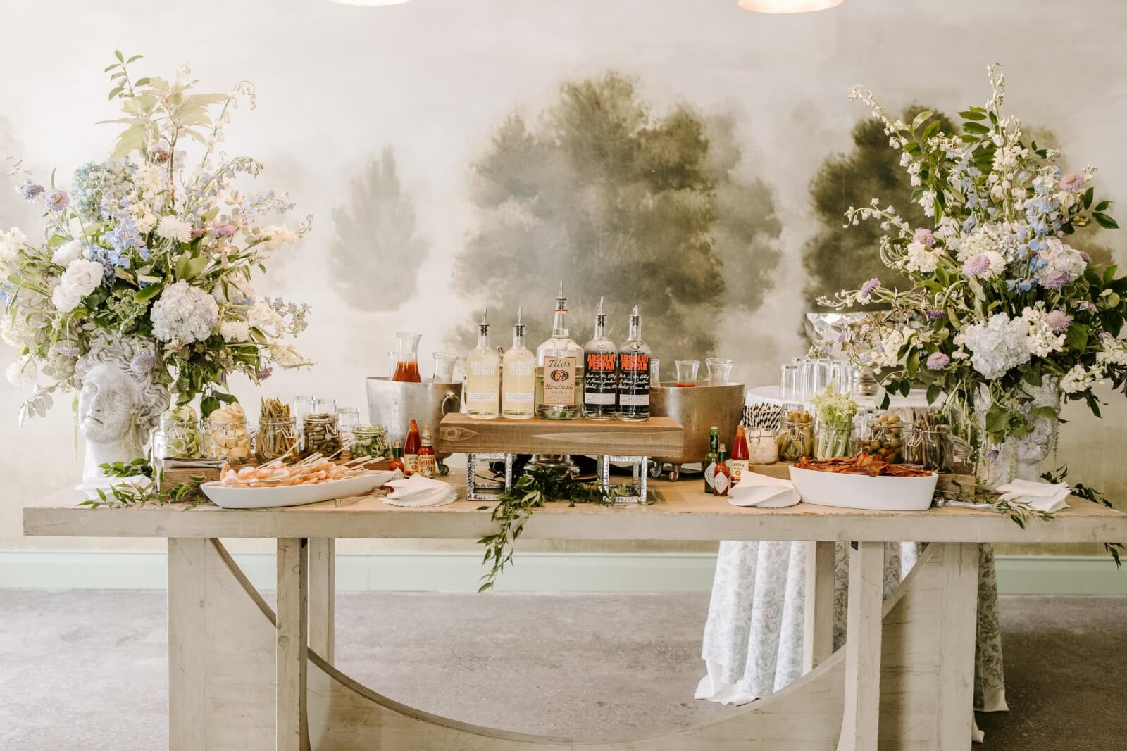 A decorated Bloody Mary bar at a Brunch Wedding in Homewood, featuring assorted food, multiple bottles of alcohol, mixers, glassware, and large floral arrangements on each side.