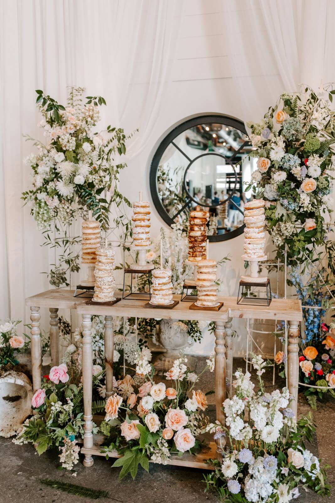 A decorated table at a Homewood brunch wedding, adorned with stacked donuts on stands and lush floral arrangements, is set beneath a round mirror.