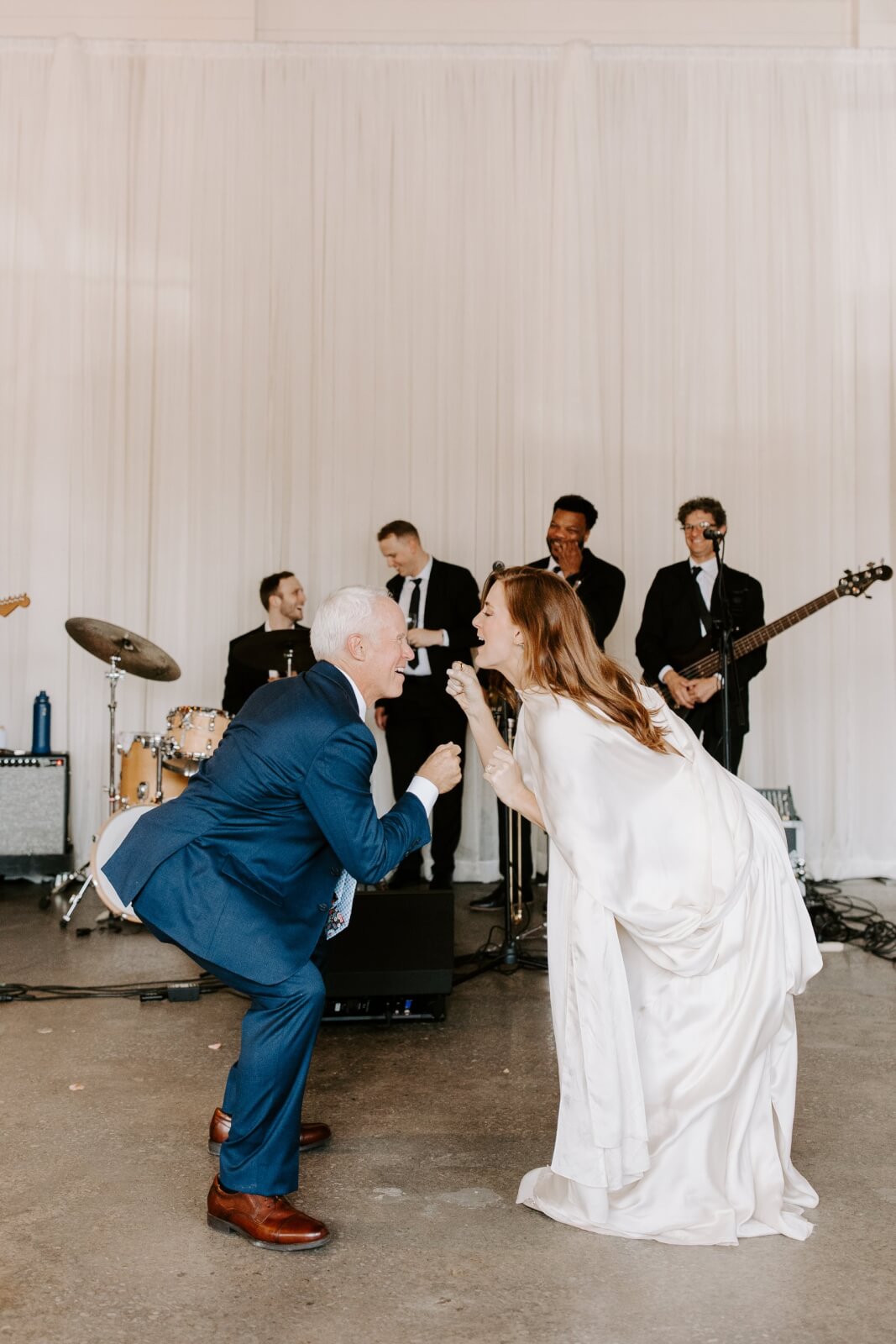 A bride and her father face each other playfully on the dance floor performing a coordinated dance while a live band performs in the background.