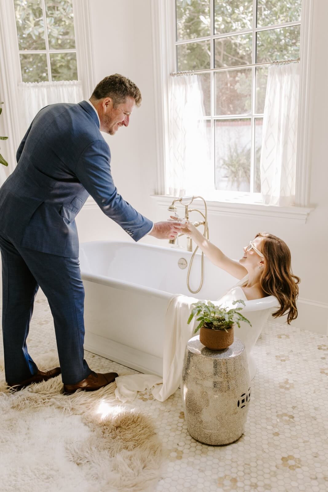 A man in a suit hands a drink to a woman relaxing in a bathtub in a bright, sunlit Homewood bathroom with large windows and a plant on a small table nearby.