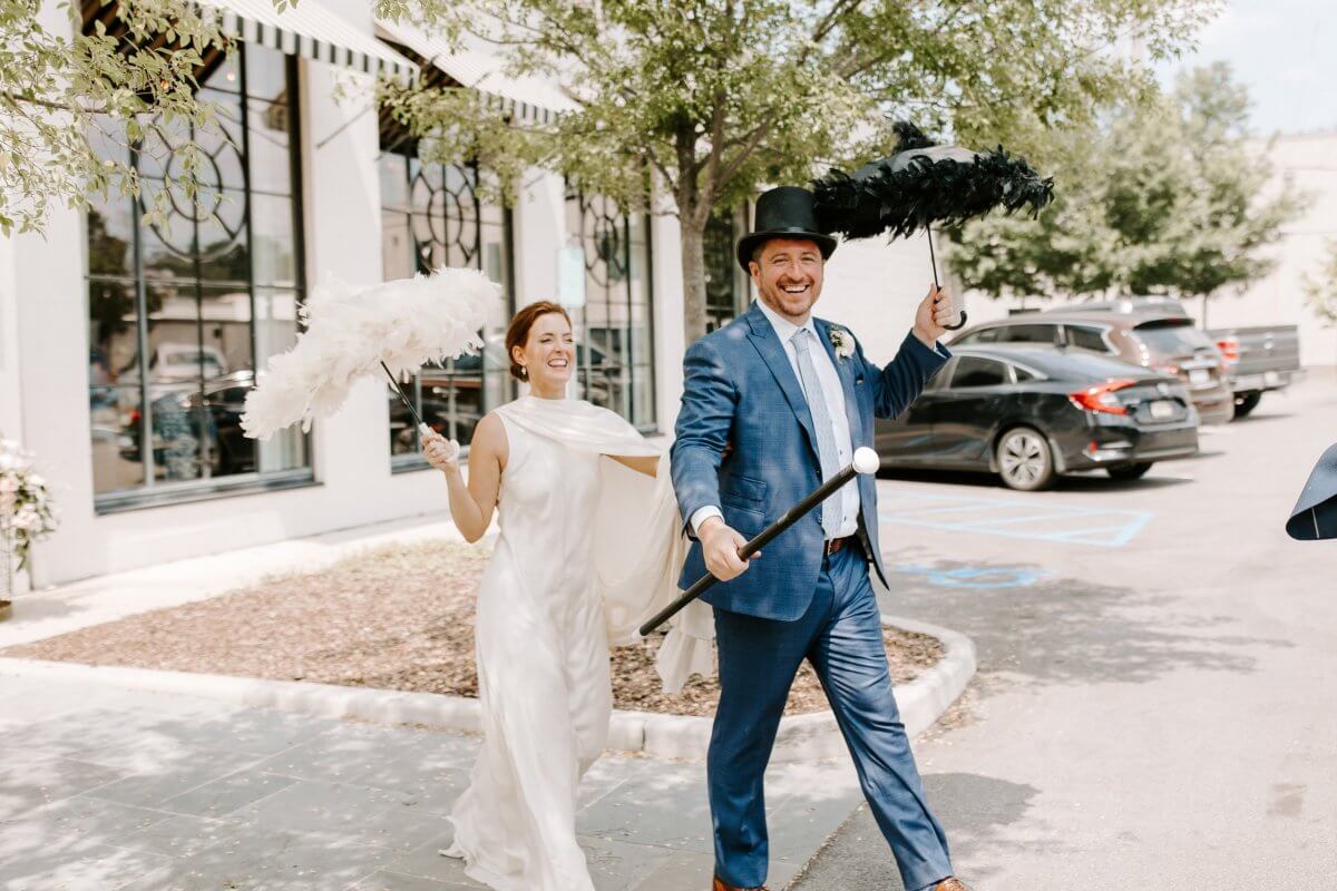 A smiling bride and groom walk outside after their Homewood wedding; the groom wears a top hat, holds a cane, and both carry feathered parasols for a festive second line sendoff.