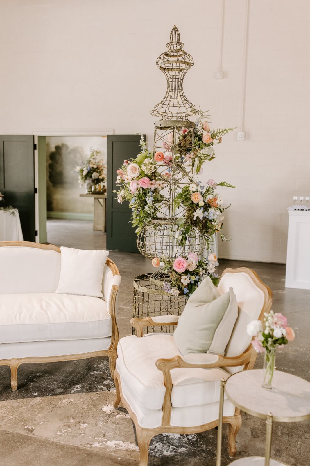 A vintage-style seating area with a white sofa, armchair, and side table sits by a tall metal floral arrangement decorated with pink and white flowers.