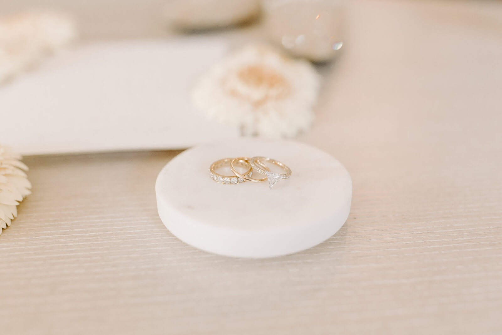 Three rings—including a triangle diamond engagement ring and two gold bands—rest on a round white marble dish.