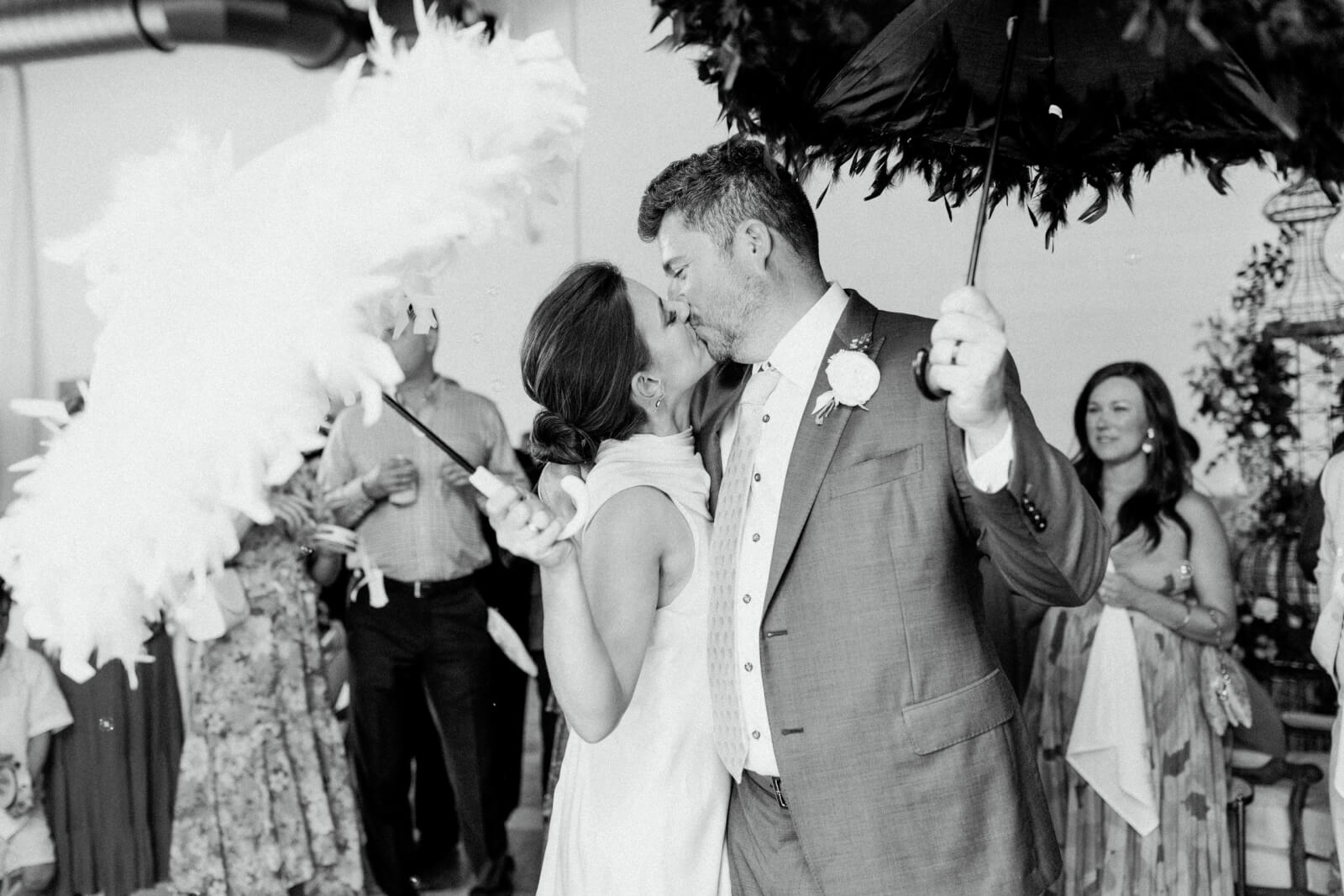 A bride and groom kiss while holding feathered umbrellas at their Homewood brunch wedding, surrounded by guests celebrating a joyful second line sendoff.