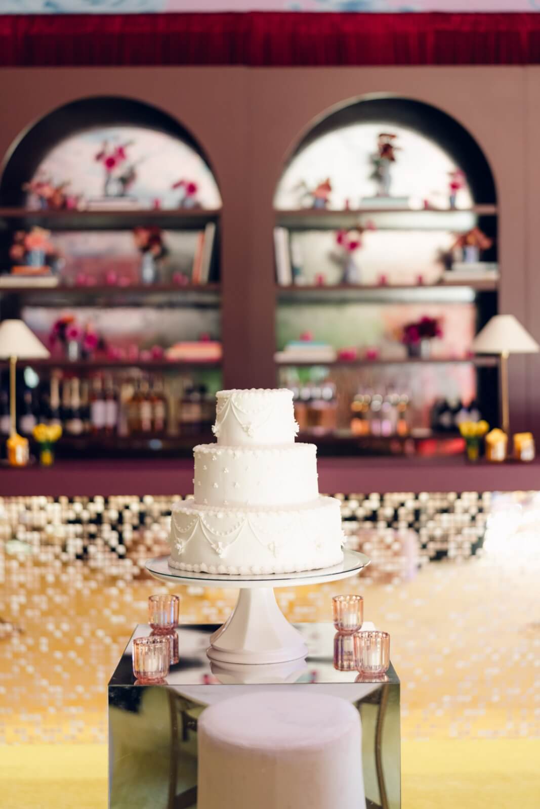 A three-tiered white cake with decorative patterns graces a stand with arched shelves behind a bar in the background.