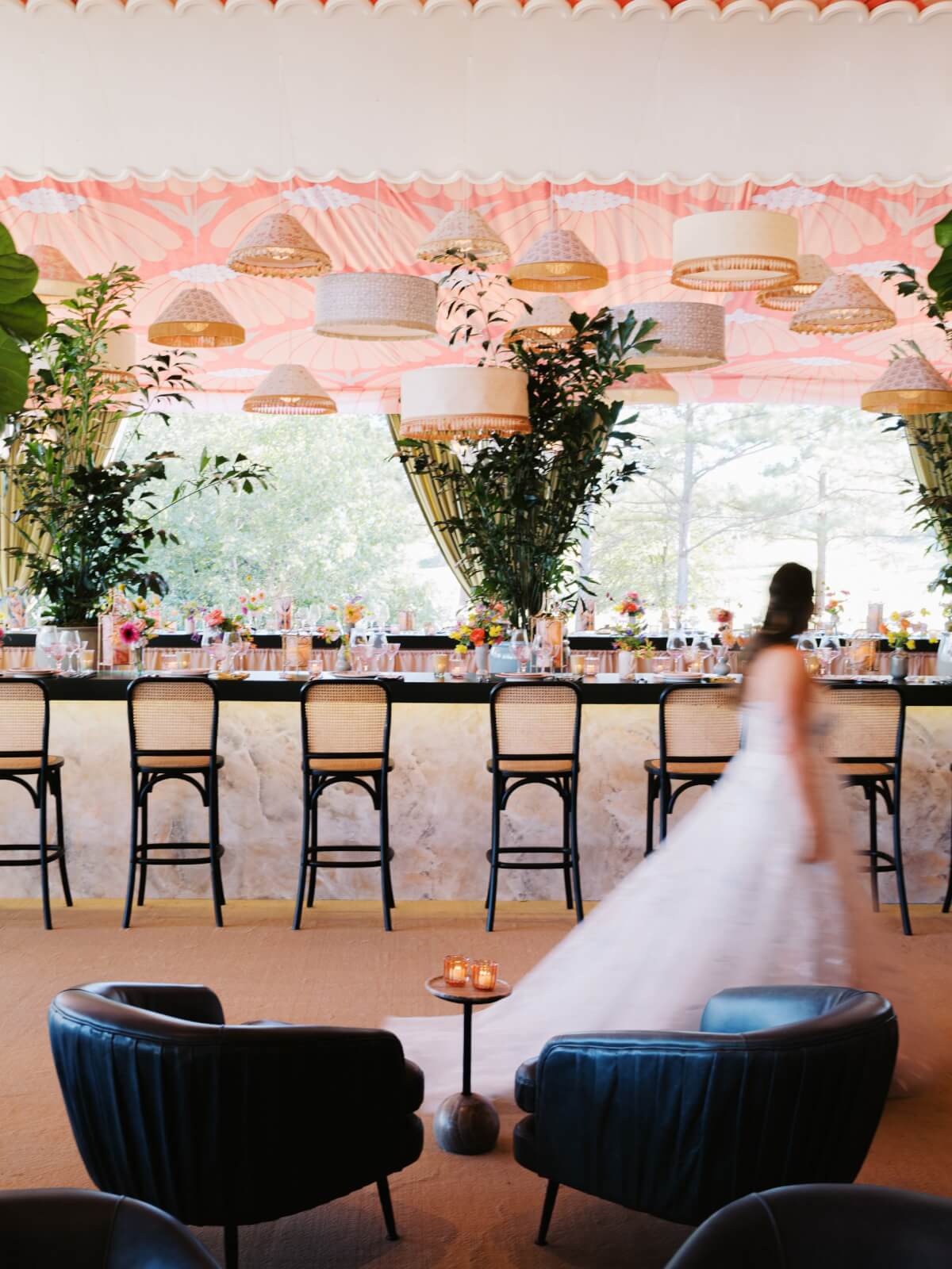 A bride in a white dress strolls past a stylish bar with high chairs, large plants, and hanging woven lamps, set against a pink and yellow patterned draped ceilng.
