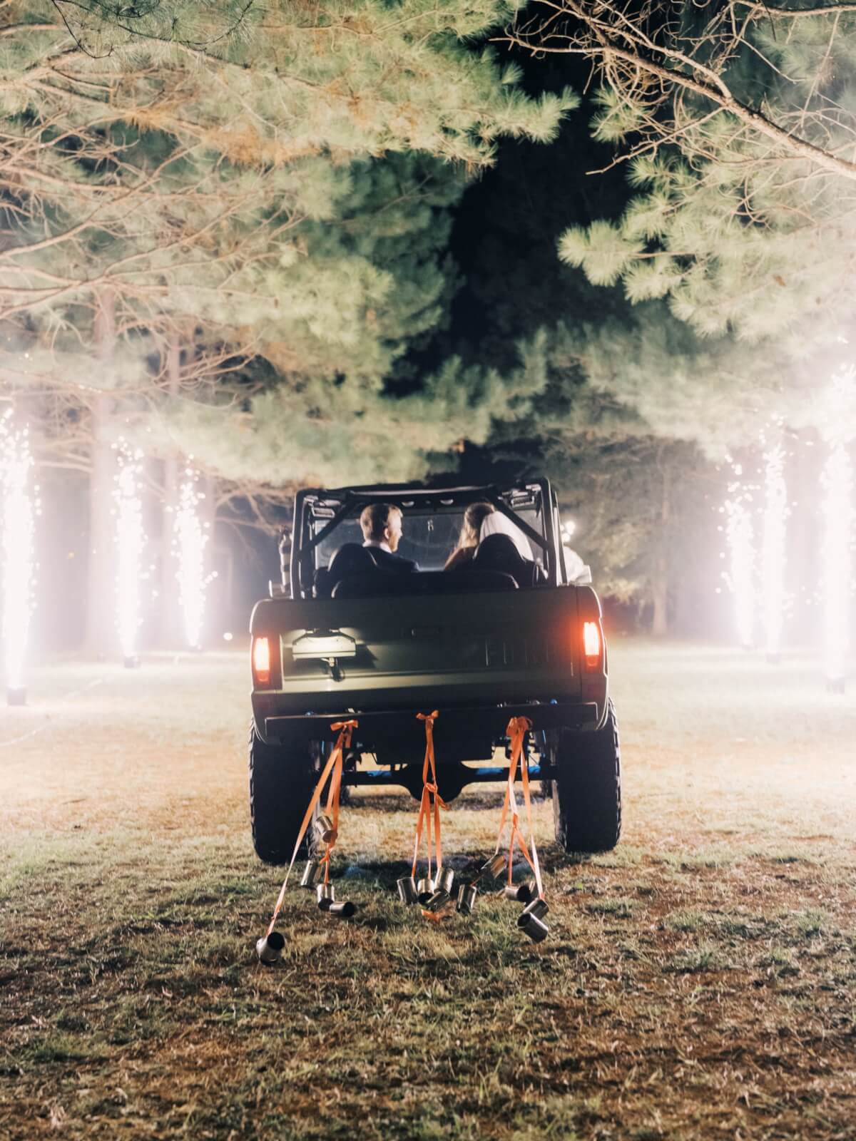 A couple drives off in an open-air Bronco under the stars, with sparklers illuminating their path and cans strung from the rear bumper. 