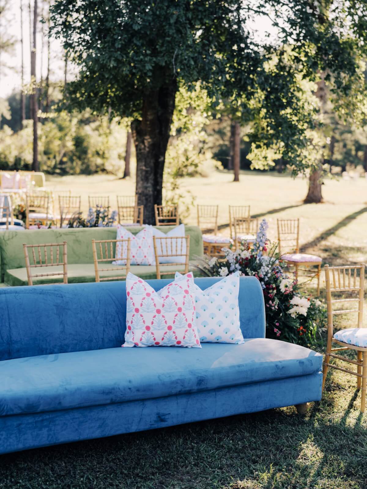 Nestled under the trees, this Outdoor Alabama Wedding setup boasts a blue sofa with patterned pillows, surrounded by wooden chairs and floral arrangements. 