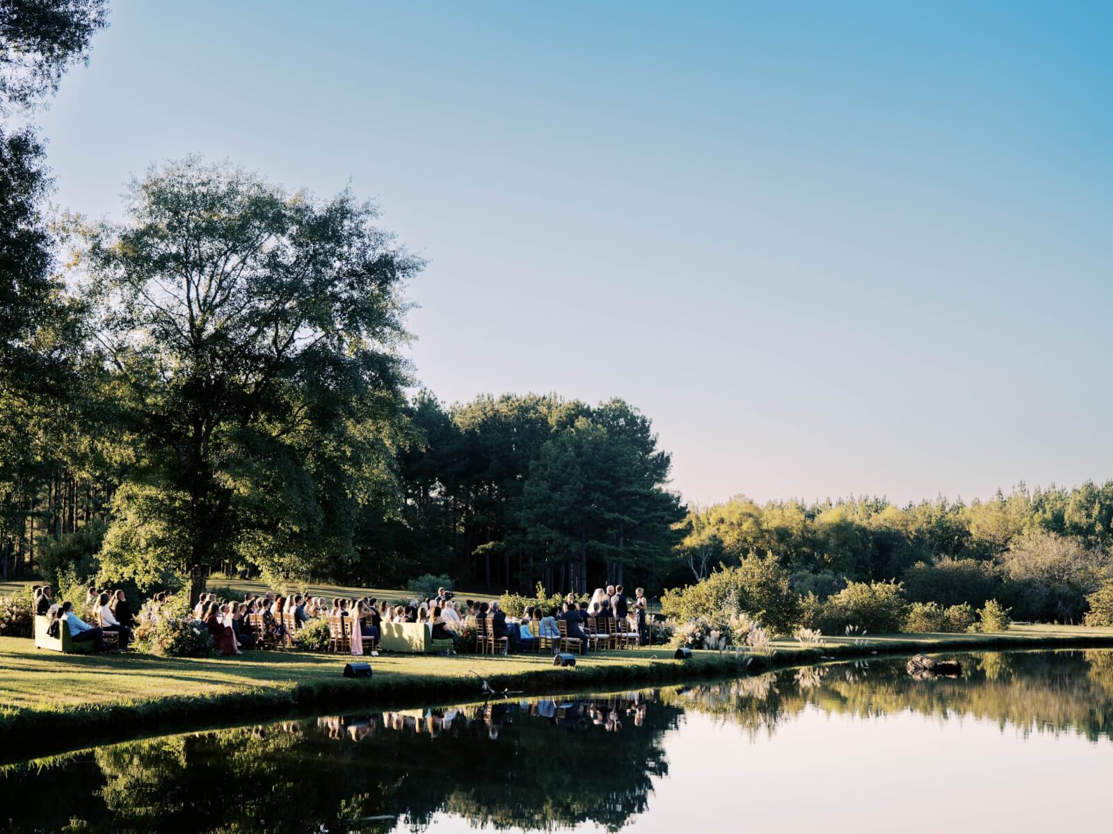 People are gathered outdoors near a lake for the wedding ceremony, surrounded by trees and greenery, with clear blue skies above.