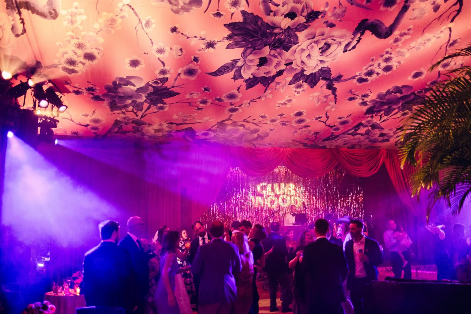 Under a floral-patterned ceiling, guests dance amidst purple lights and a "Club Wood" backdrop. 