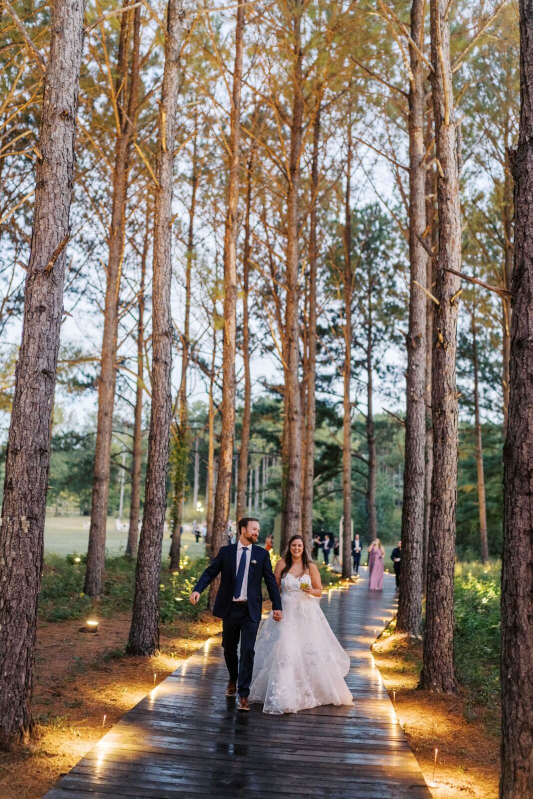 A couple in wedding attire walks hand in hand down a wooden path lined with trees and lights.