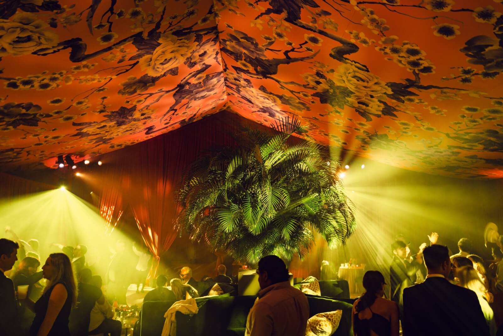 At a dimly lit reception dance floor guests mingle beneath an ornate floral ceiling, as bright yellow spotlights illuminate the room. A large green plant adds to the ambiance curated by a world-class event designer.