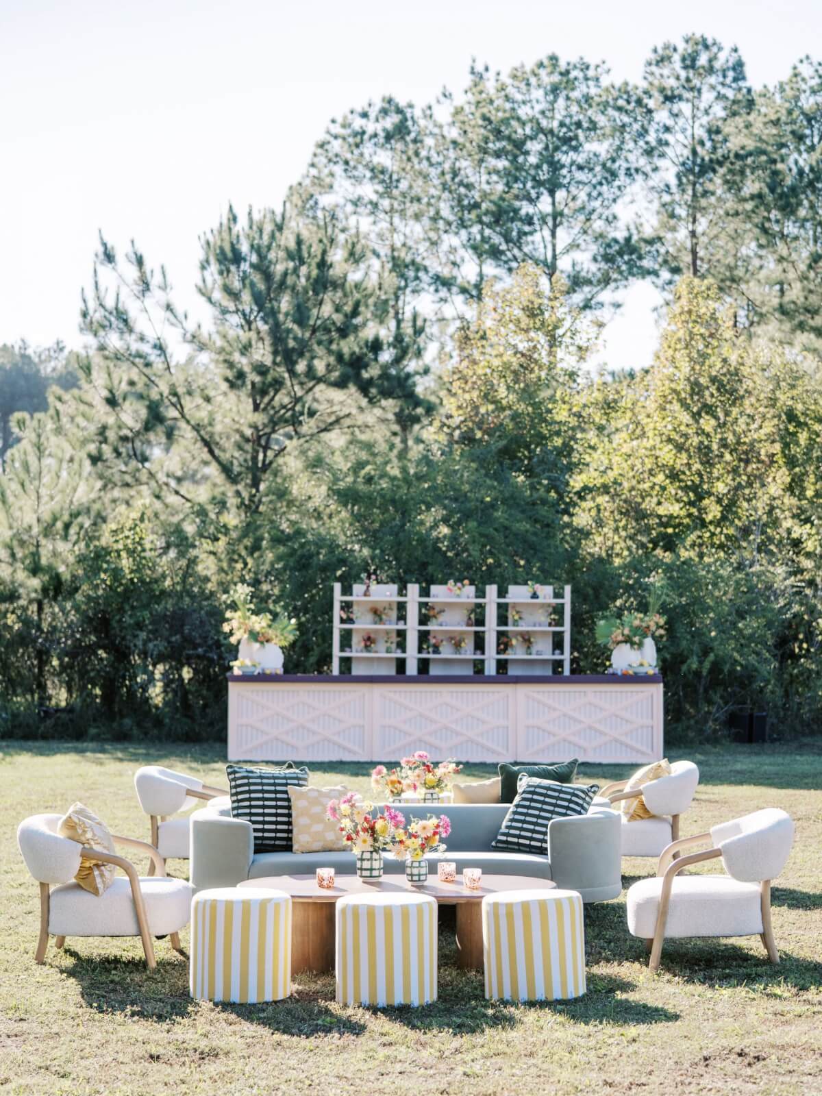 An outdoor cocktail hour in an open field features a walk-up bar with elegant seating in the foreground.
