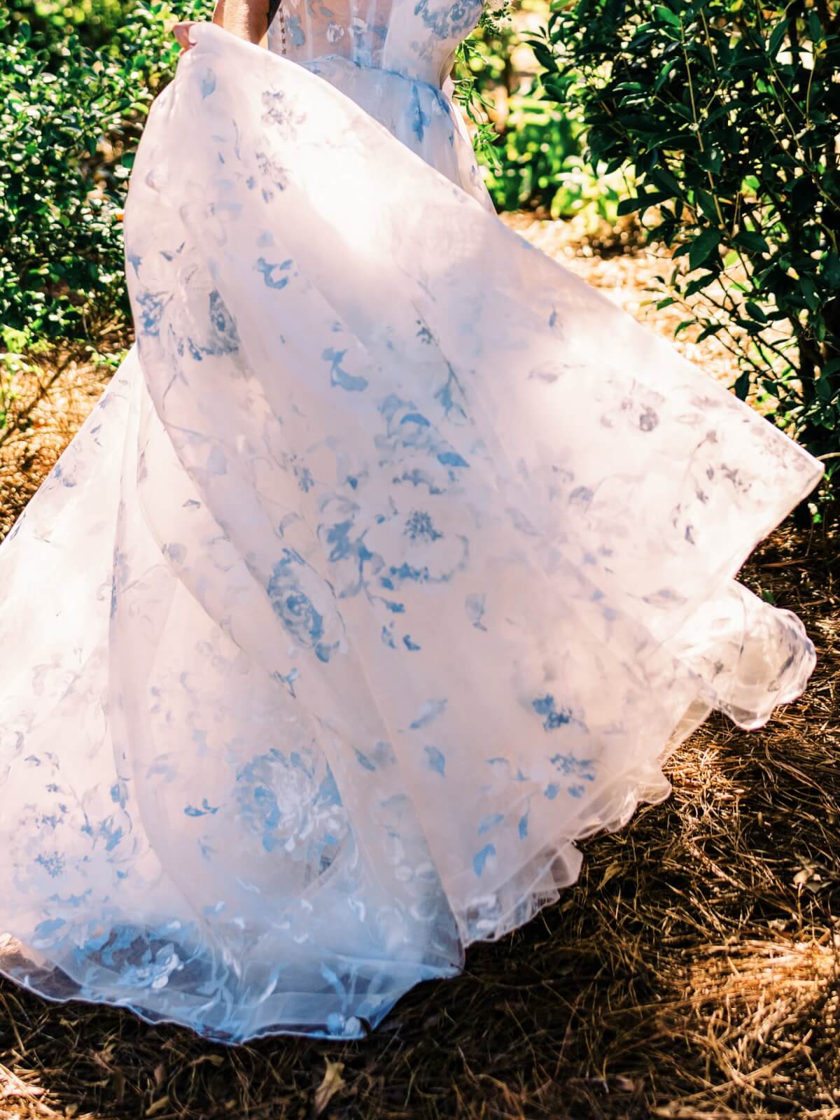 A bride in a flowing, white gown with blue floral-patterns twirls outdoors near lush green bushes.