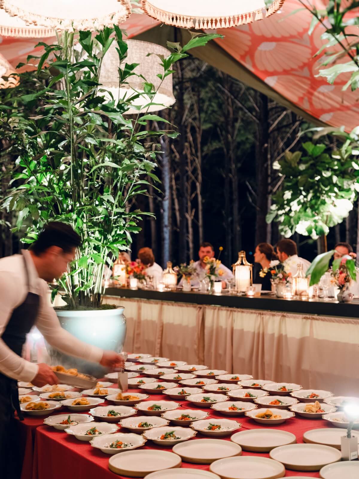 A server prepares plates of food on a red tablecloth. Guests sit at a long table adorned with lanterns and flowers, surrounded by lush greenery.
