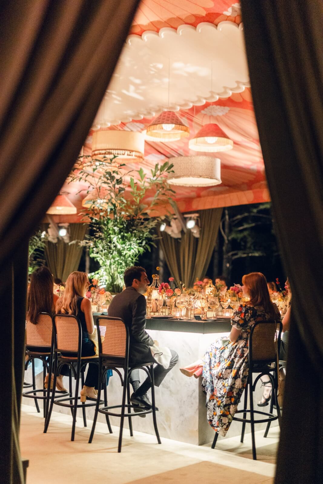 People sitting at a bar under a tent with warm lighting, surrounded by plants.