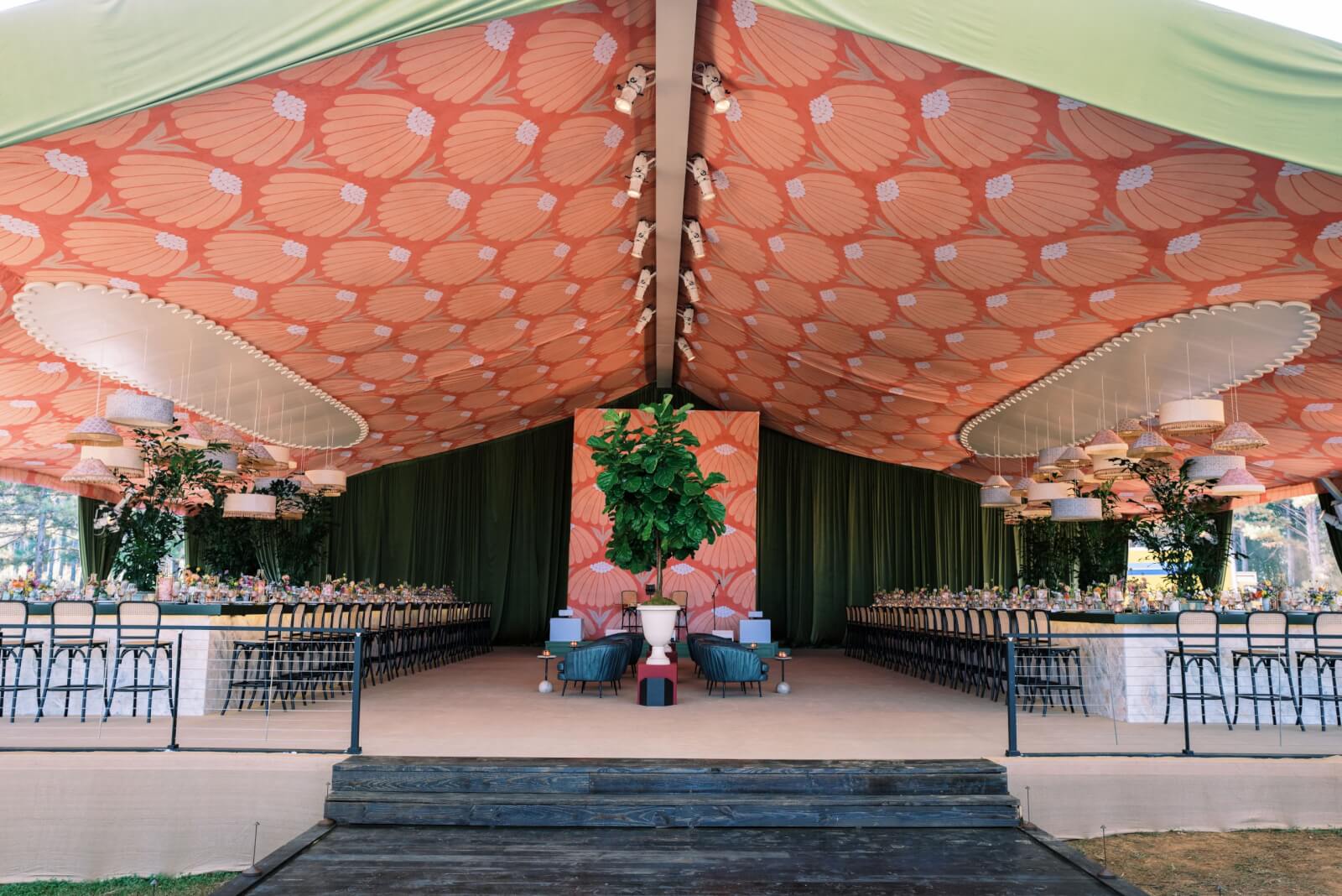 An ornate tent with a symmetrical design, showcasing two long dining tables, a patterned ceiling, hanging lights, and greenery.