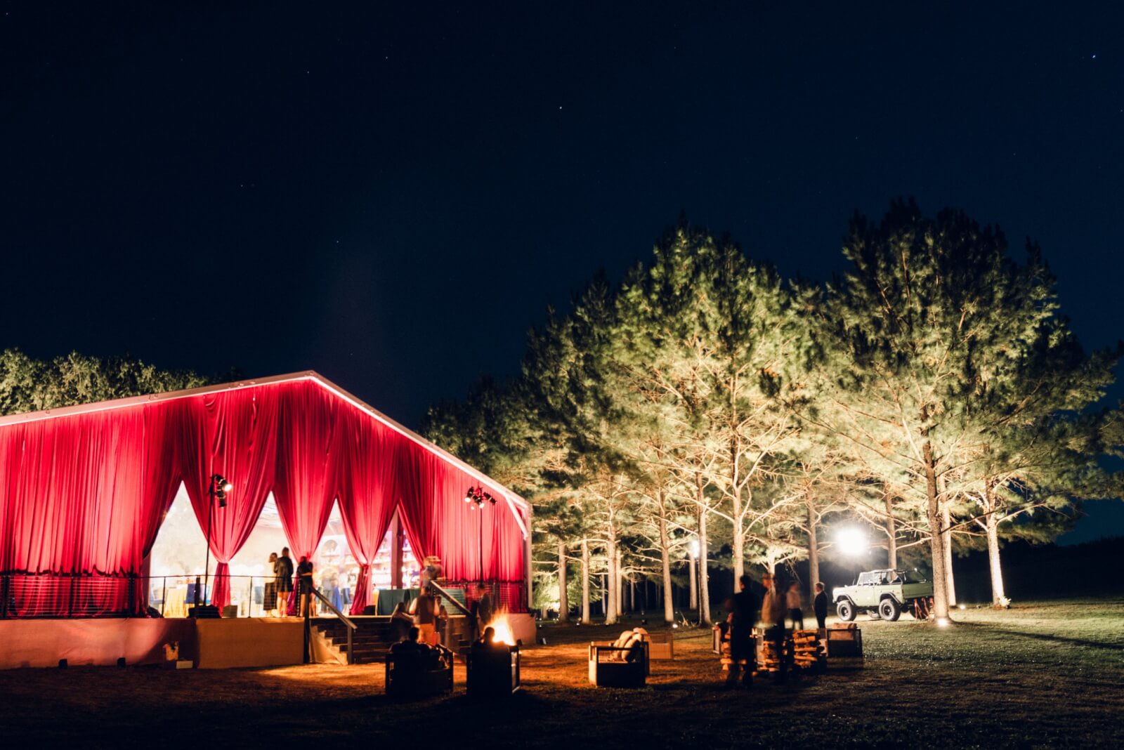 A red-draped tent lit up at night next to illuminated trees creates a stunning backdrop for an outdoor Alabama wedding. A green Bronco is parked nearby, and people are gathered outside.