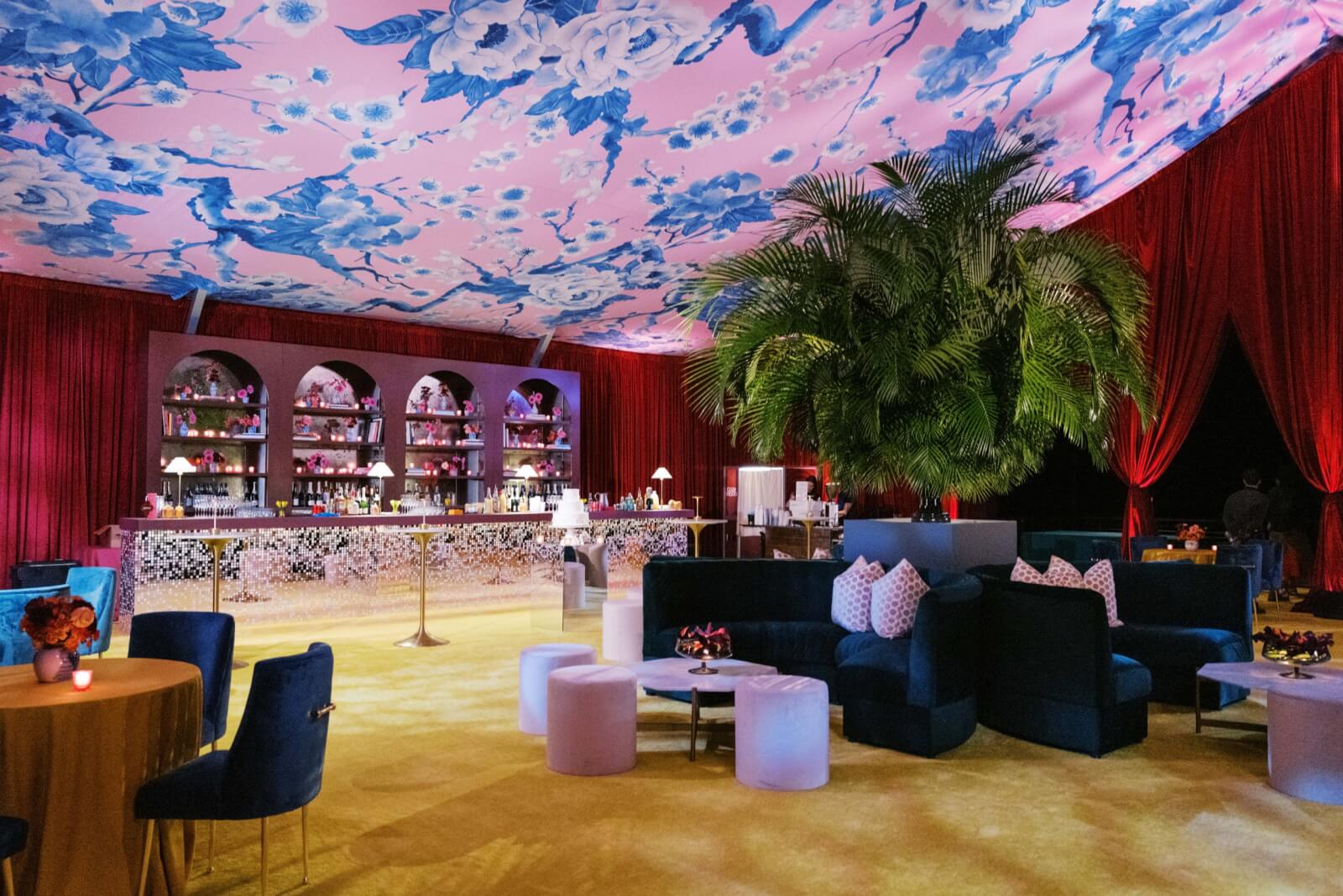 Luxurious lounge with floral ceiling, velvet furniture, and red curtains. A decorative tree stands in the middle of the space with lounge chairs surrounding it. 