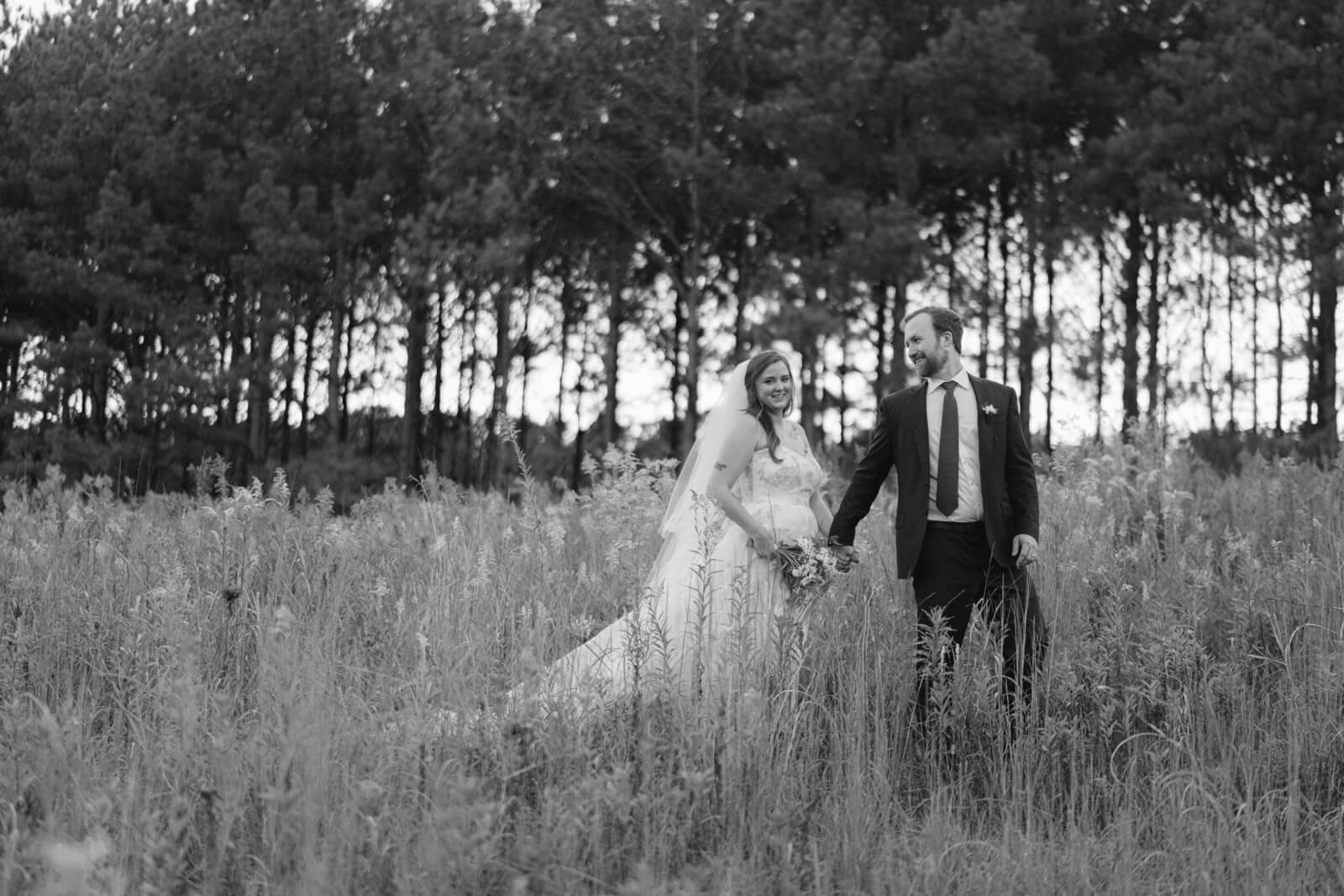A black and white photo of a couple in wedding attire strolls hand in hand through a grassy field, tall trees framing the scene. 