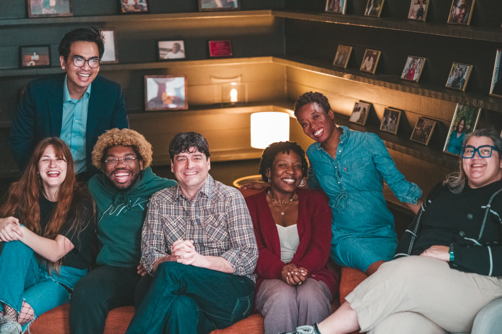 The Create Birmingham Staff (Quang Do, (president and CEO), Madi Vordenbaum, Desean Motley, Lee Shook, Imani Richardson, Viola Ratcliffe, and Jessica Moody), are sitting and standing in a cozy room with a lit lamp and photos on shelves, smiling at the camera.