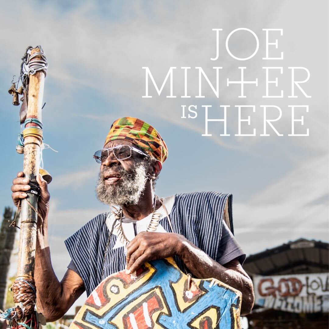 Joe Minter, an elderly man with a beard, wearing a colorful hat and traditional clothing, holds a staff as he stands before a vibrant handmade sign. "Joe Minter is Here" overlays the clouded sky.