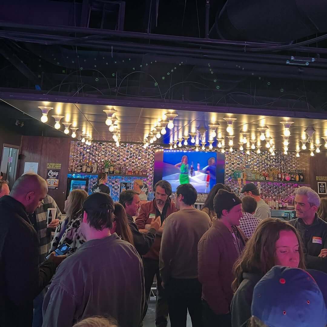 A bustling bar buzzes with people chatting and checking their phones, while the ceiling glimmers with lights. A large screen showcases two figures on stage.